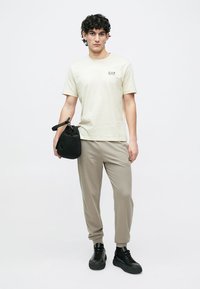 Lys beige t-skjorte med EA7-logo, kombinert med lysegrå joggebukser og svarte joggesko. Modellen holder en liten svart veske.