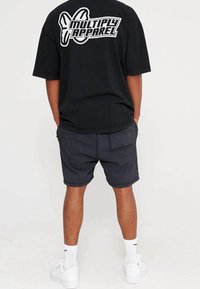 Černé oversized tričko s bílým logem "MULTIPLY APPAREL" na zadní straně, spárované s tmavými šortkami a bílými teniskami. Neformální design oblečení.
