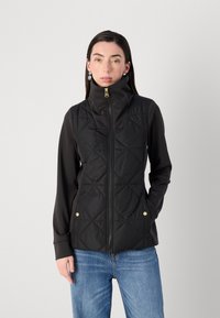 Een vrouw staat zelfverzekerd, gekleed in een zwart gewatteerd vest met een hoge kraag, gecombineerd met een lange mouwen top en blauwe jeans, tegen een neutrale achtergrond.
