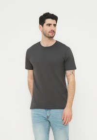 Άνδρας με σκούρα μαλλιά και τατουάζ στο αριστερό χέρι, φορώντας απλό σκούρο γκρι μπλουζάκι και ανοιχτό μπλε τζιν, κοιτάζοντας προς τα δεξιά.