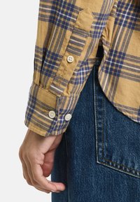 Gul og blå ternet skjorte med lange ærmer og knapmanchetter samt et broderet logo. Parret med mørk denim jeans.