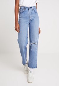 Vaqueros ajustados de color azul claro con un desgarro en la rodilla, combinados con calcetines blancos y zapatillas blancas, junto con una camisa blanca metida por dentro.