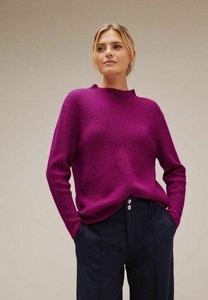 Leuchtend magentafarbener Strickpullover mit hohem Kragen, der strukturierte Muster und einen lässigen Schnitt aufweist. Kombiniert mit maßgeschneiderten, dunklen Hosen.