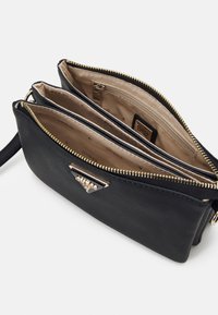 Černá taška z umělé kůže se zlatým kováním, vybavená dvěma kapsami na zip a béžovou textilní podšívkou s potiskem názvu značky uvnitř.