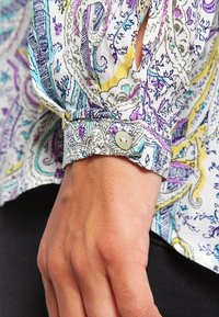 Camicia a maniche lunghe con un motivo paisley colorato. Presenta un polsino con bottoni e un tessuto morbido e leggero. Predominantemente bianca con accenti viola, blu e gialli.