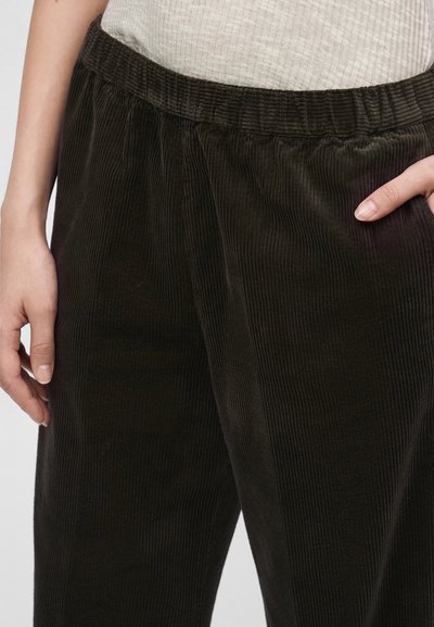 Gros plan d'une personne portant un pantalon en velours côtelé noir avec une ceinture élastique et un haut côtelé de couleur claire, les mains détendues le long du corps.