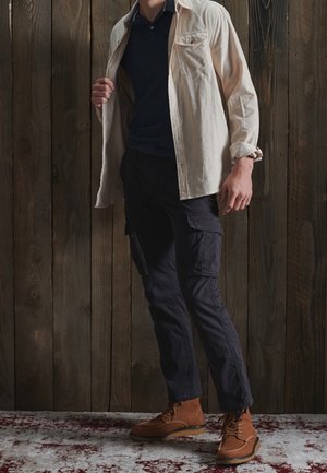 Homme portant une veste beige ouverte sur une chemise noire, un pantalon cargo noir et des bottes marron à lacets, debout sur un tapis contre un mur en bois.