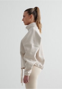 Béžová mikina na zip s vysokým límcem, volným střihem a elastickými manžetami. Šňůrky na spodním lemu přidávají nastavitelné prvky.