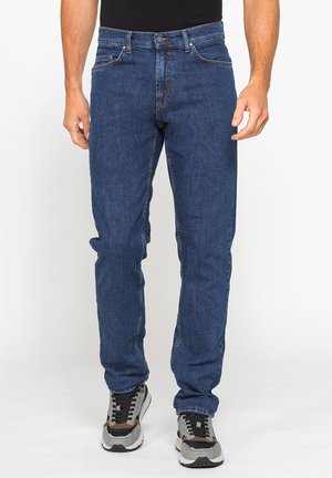 Uomo che indossa jeans blu a gamba dritta e scarpe da corsa grigie, in piedi davanti a uno sfondo neutro.