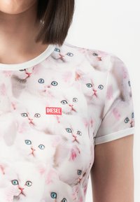 Kvinde iført en hvid T-shirt dækket med flere hvide katteansigtter med blå øjne og et lille rødt Diesel-logo på brystet.