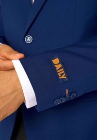 Vīrietis pielāgo tumši zilā uzvalka piedurkni ar oranžo "DIENAS" etiķeti, zem tā valkājot baltu krekla, un piedurknes zilajiem pogām.