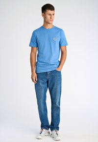 Ljusblå bomulls T-shirt med kort ärm och rund halsringning, med en liten solrosgrafik på bröstet. Bärs med blå denimjeans.