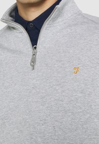 Felpa gray con zip realizzata in tessuto morbido, caratterizzata da un colletto alto e una camicia blu navy sotto. Logo giallo piccolo e distintivo sul petto.