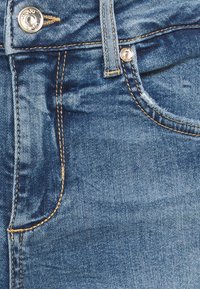 Pantalones vaqueros de mezclilla en azul medio con costuras doradas, que cuentan con un bolsillo delantero, botón de metal y cierre de cremallera. Tejido texturizado.