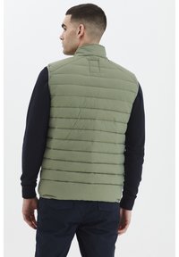 Svijetlozeleni puff jacket s horizontalnim šavovima i visokim ovratnikom, nosi se preko tamnoplave košulje dugih rukava, prikazan s leđa.