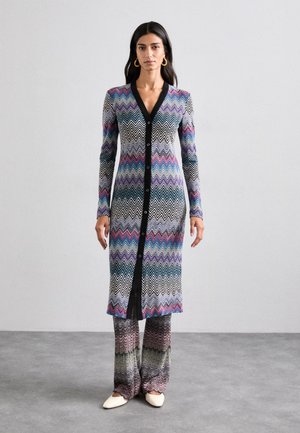 Long knit cardigan with a black v-neckline, featuring a multicolored zigzag pattern. Paired with wide-leg pants in matching colors and a subtle sheen.
