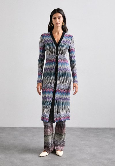 Cardigan long en tricot avec un col en V noir, arborant un motif zigzag multicolore. Assorti à un pantalon ample dans des couleurs coordonnées et avec un subtil éclat.