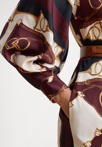 Rochie de mătase cu un imprimeu îndrăzneț în culoarea bordo și crem, cu motive ecvestre aurii. Mâneci lungi cu umeri adunați și un accent cu curea maro.