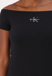 Juodas medvilnės viršus be petnešų, su sidabriniu logotipu ir užrašu "Calvin Klein" virš stilizuoto "CK" priekyje.