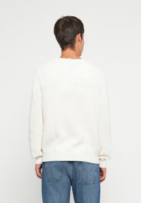 Balts adīts džemperis ar ribotām faktūrām un nokritušiem pleciem, ar apaļu kakla izgriezumu un garām piedurknēm, kombinēts ar gaiši ziliem džinsiem.