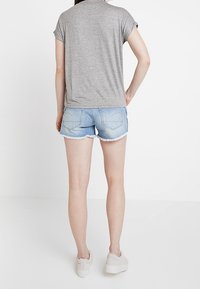 Camiseta gris de manga corta, pantalones cortos de mezclilla con dobladillo deshilachado, combinados con zapatillas blancas de plataforma; vista desde la espalda, sobre un fondo blanco liso.