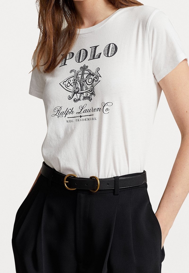 Femme portant un t-shirt blanc Ralph Lauren Polo avec logo, rentré dans un pantalon noir taille haute, ceinturé d'une bande noire avec une boucle dorée.