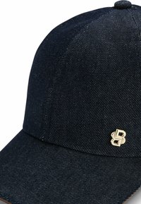 Mörk denimkeps med låg skärm. Har en guldlogotyp på framsidan. Texturerat tyg med kontrasterande sömdetaljer.