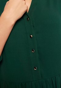 Zielona bluzka z tkaniny, z dekoltem w serek, ozdobiona złotymi guzikami i krótkimi rękawami. Marszczenia u dołu dodają faktury.