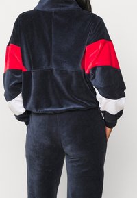 Marineblauw velours trainingspak met een hoge kraag. Voorzien van rode en witte horizontale strepen op de mouwen, een zachte textuur en een comfortabele pasvorm.