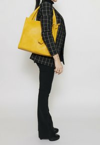 Geel lederen tote bag met een textuurafwerking en dubbele handvatten, vastgehouden aan de zijkant. Draagt een zwart-witte blazer met ruitpatroon en flared broek.