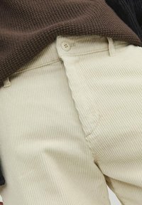 Creme corduroy broek met verticale ribbels, een knoopsluiting en zoomstiksel, gecombineerd met een bruine gebreide trui.