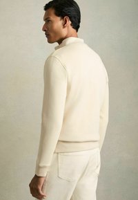 Un pulover crem cu mâneci lungi, guler înalt și manșete ribbed, purtat cu pantaloni bej deschis, evidențiind o țesătură netedă și o croială modernă.