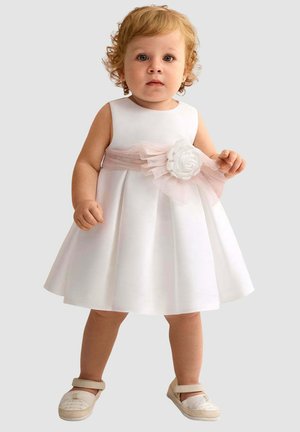 Bambino con capelli ricci che indossa un vestito bianco senza maniche con una cintura rosa e un fiore, in piedi con scarpe beige su uno sfondo semplice.