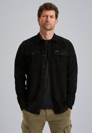 Zwarte flanellen overhemd met een geruit patroon, twee borstzakken en knoopsluiting. Draagt over een zwart shirt. Olijfgroene cargobroek zichtbaar.