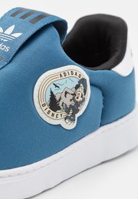 Blue fabric sneaker with white rubber sole. Features a round Disney Adidas patch depicting a cartoon character in an outdoor scene.