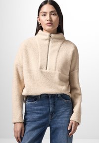 Pullover in pile beige con colletto alto, apertura frontale a metà zip e una texture morbida. Abbinato a jeans in denim blu con leggeri segni di usura.