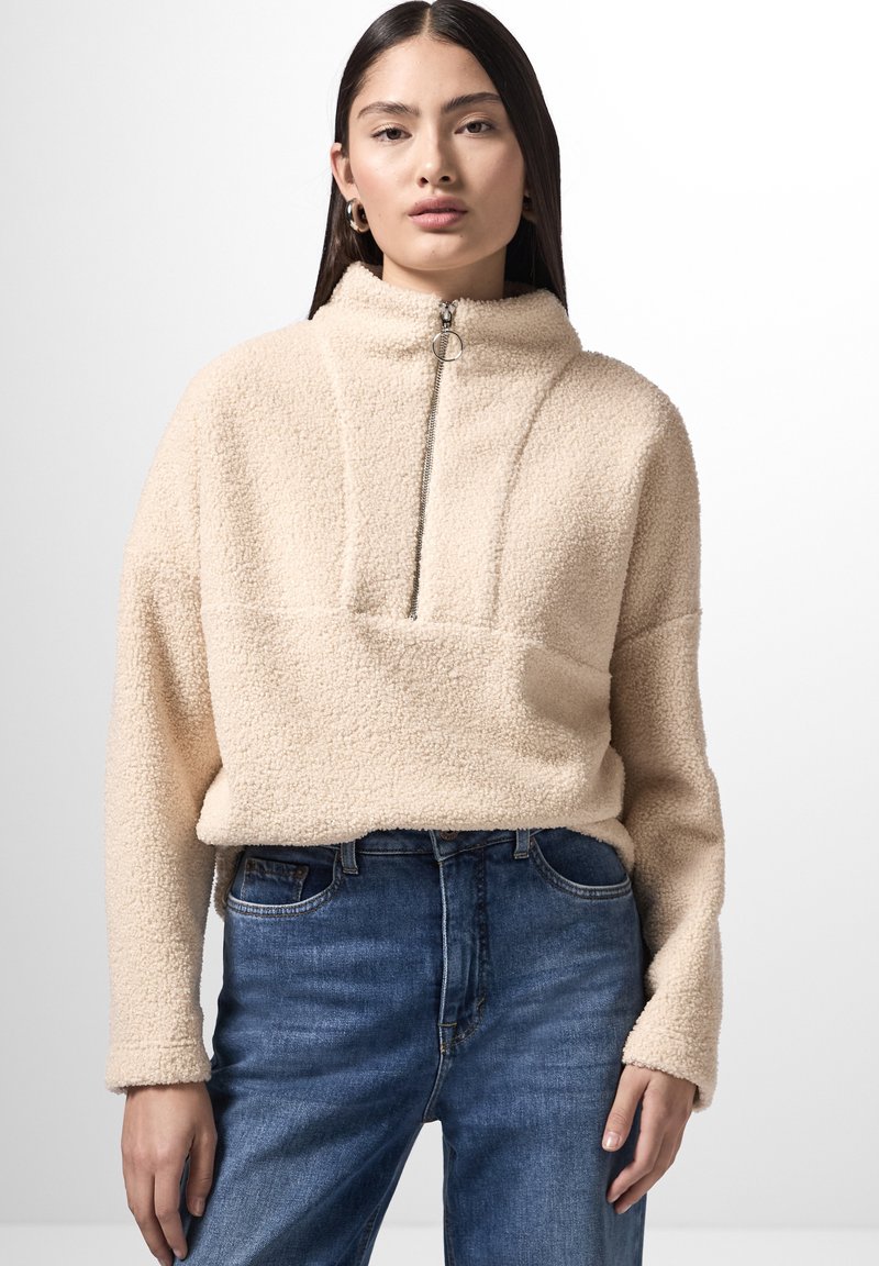 Pullover in pile beige con colletto alto, apertura frontale a metà zip e una texture morbida. Abbinato a jeans in denim blu con leggeri segni di usura.