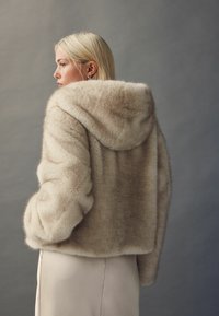 Žena s krátkymi blond vlasmi, oblečená v béžovom kožuchu s kapucňou, pohľad zozadu na jednoduchej šedej pozadí.