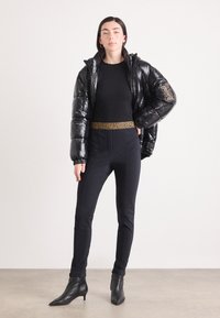 Doudoune noire en matériau brillant, avec une ceinture à motif léopard et un pantalon noir slim. Le modèle porte des bottines noires à la cheville.