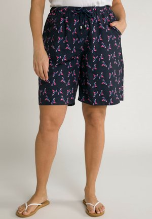 Short bleu marine avec motif d'oiseaux roses et bleus, taille élastique avec cordon, poches latérales, porté avec des tongs blanches et un haut blanc.