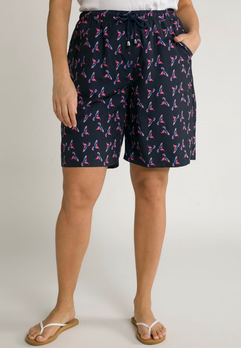 Short bleu marine avec motif d'oiseaux roses et bleus, taille élastique avec cordon, poches latérales, porté avec des tongs blanches et un haut blanc.