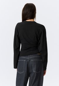 Haut noir à manches longues avec un design ajusté, présentant un détail de dos torsadé. Associé à un jean en denim bleu foncé avec des coutures contrastantes.