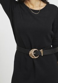 Cintura in pelle nera con una fibbia a forma di conchiglia in tono oro, caratterizzata da dettagli curvi e un aspetto testurizzato, indossata sopra un indumento nero.