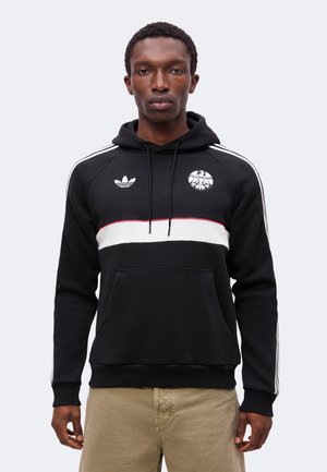 Homme portant un sweat à capuche noir avec une bande blanche et rouge, logo Adidas et Eintracht, associé à un pantalon beige, regardant vers l'avant.