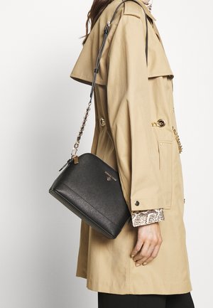 Sac bandoulière noir en simili cuir avec fermeture éclair, ferrures dorées et finition texturée, porté avec une chaîne sur un manteau beige.