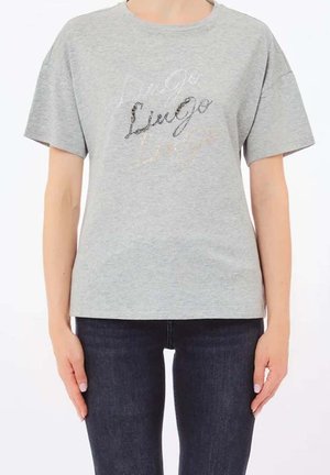 Persona con camiseta gris claro de manga corta con texto brillante y vaqueros oscuros, mostrada desde los hombros hasta las caderas sobre un fondo liso.