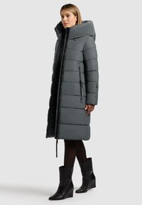 Pufferjakke i mørkegrå, med høj krave, lynlås og sidelommer. Glat tekstur med quiltet design. Bæret med sorte hælboot.