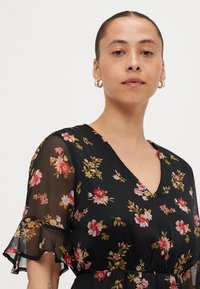 Robe noire à fleurs avec de courtes manches volantes. Présente un décolleté en V et des motifs floraux verticaux en rose, jaune et vert sur un tissu transparent.