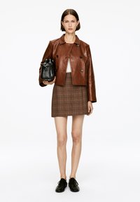 Braune Leder-Shortjacke mit doppelten Knöpfen, weißes Oberteil, karierten Minirock in Braun und Creme sowie schwarze Handtasche. Schwarze Schnürschuhe.