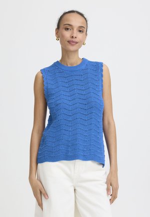 Femme portant un haut tricoté bleu sans manches et un pantalon blanc, debout devant un fond clair uni, regardant la caméra.